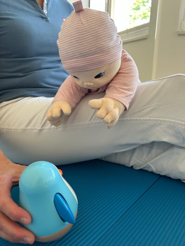 Baby-Puppe in Bauchlage über dem Oberschenkel, greift nach einem Spielzeug – demonstriert eine therapeutische Übung zur Stärkung der Nacken- und Rumpfmuskulatur