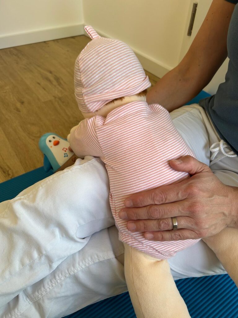 Seitenansicht der Bauchlage über dem Oberschenkel mit unterstützender Hand – gezeigte Haltung fördert die Kopfkontrolle und Gleichgewichtsentwicklung