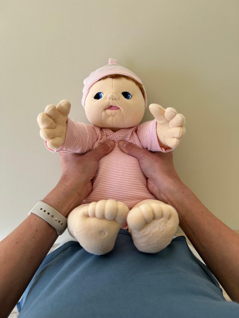 Therapeutisches Hochnehmen eines Babys in Rückenlage mit Griff am Schultergürtel – Demonstration mit Puppe in der Kinderphysiotherapie Ritterburg